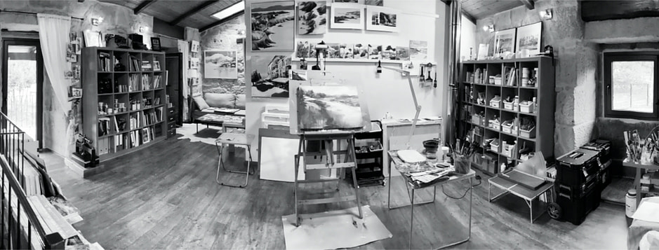 A peek into the artist studio with a painting wall in front, art supply shelving on the right and left and a cozy space for a chat with clients on the left back.